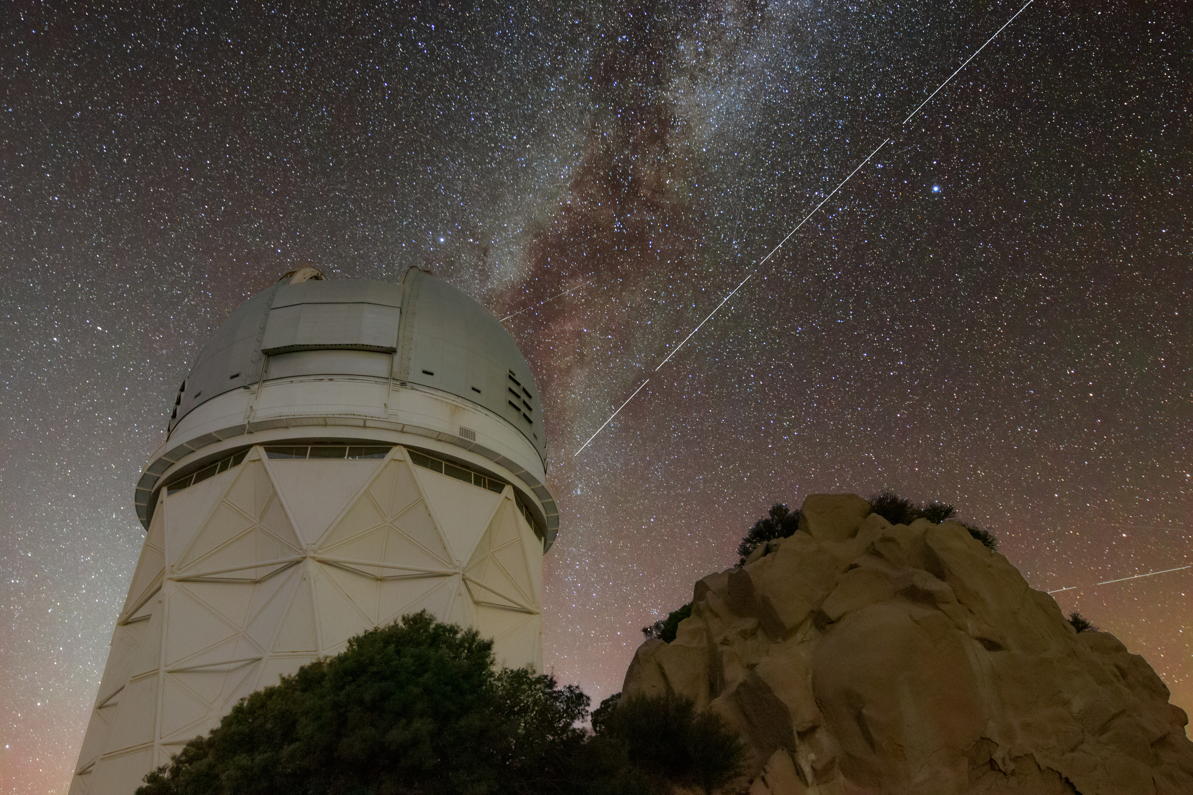 Foreground: Nicholas U. Mayall 4-meter Telescope at Kitt Peak National Observatory in Arizona. Background: the night sky with the Milky Way stretching across the center of the frame. A bright streak from the BlueWalker3 satellite cuts across the night sky.