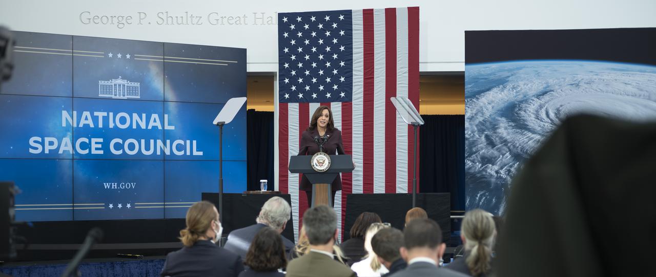 Vice President Kamala Harris speaking on a small stage during a National Space Council meeting. Behind here is a large American flag, and to either side of here are large screens. One depicts the logo for the National Space Council, while the other shows an image of the Earth as taken from space.